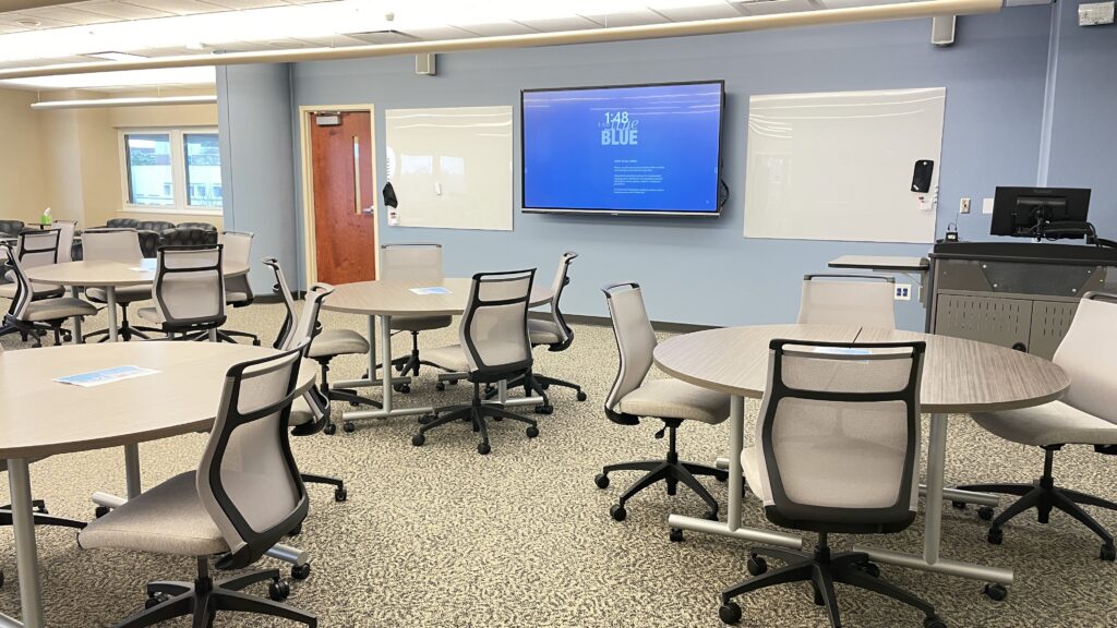 A modern meeting room with round tables and mesh chairs, a large display screen at the front, and two whiteboards mounted on the wall. A computer workstation is near the display.