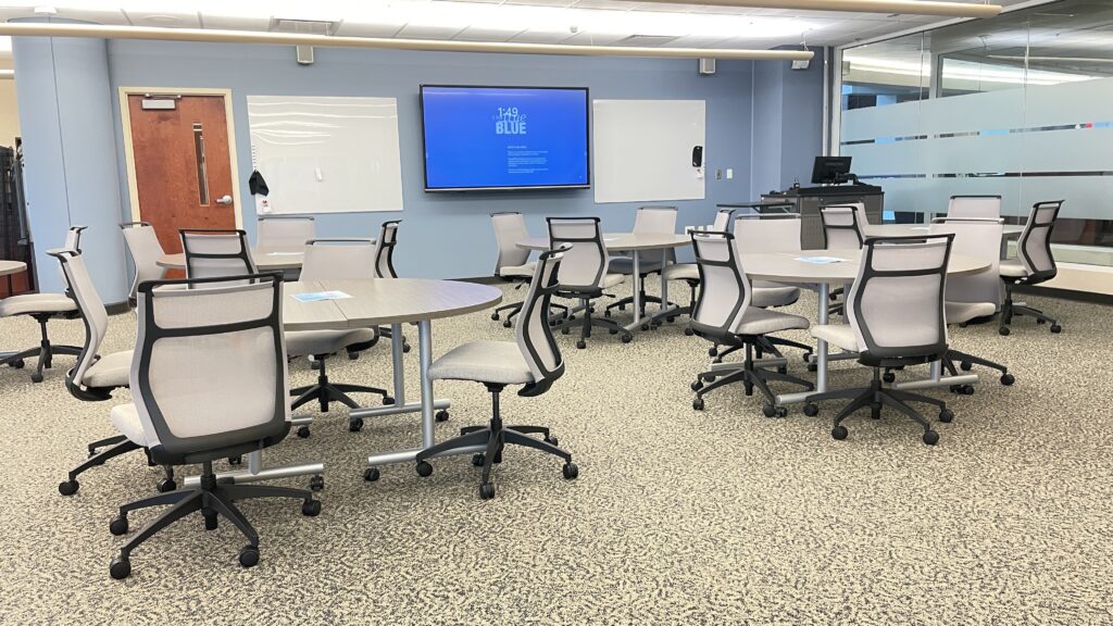 The CTM event space features several round tables with mesh chairs, a large wall‑mounted display screen, whiteboards on either side of the display, and a computer workstation.
