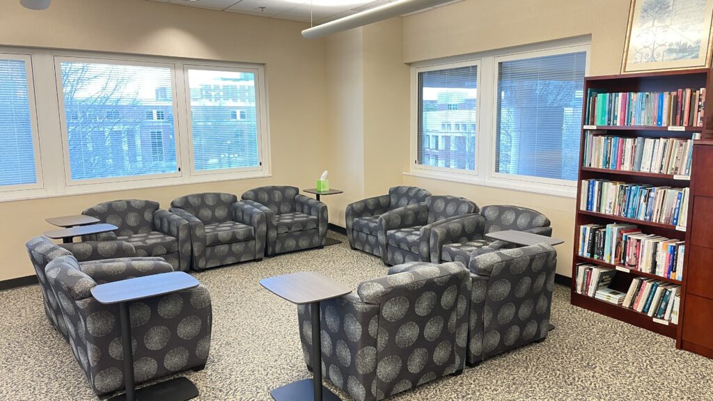 A soft‑seating area with patterned armchairs arranged in a circular layout around small side tables. Multiple windows provide natural light, and a bookshelf stands along one wall.